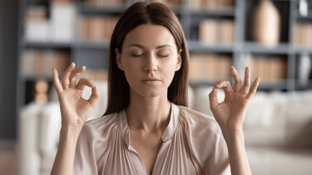 Woman sitting in a library with her eyes closed, thumbs and index fingers joined on both hands, meditating.