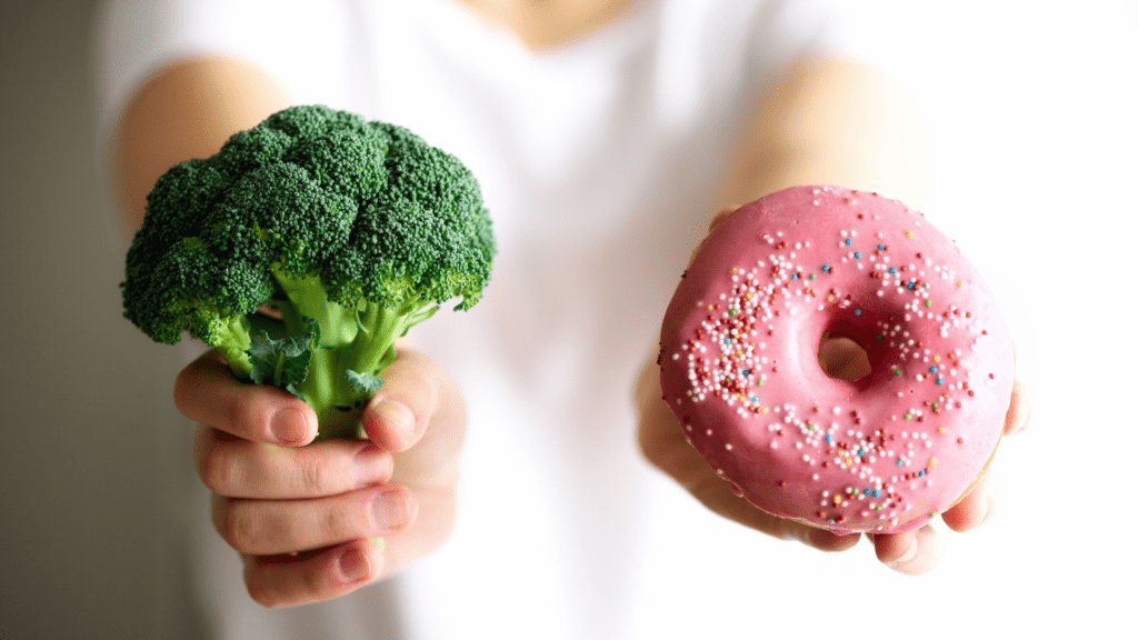 A person holding broccoli in one hand and a pink frosted doughnut in the other hand.