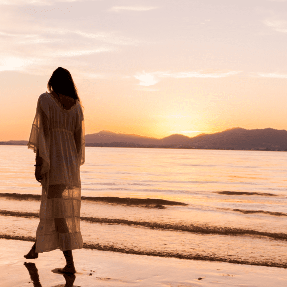 Woman walking barefoot along a sandy beach at sunset.