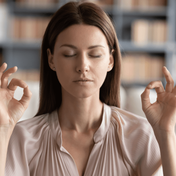 Woman sitting in a library with her eyes closed, thumbs and index fingers joined on both hands, meditating.